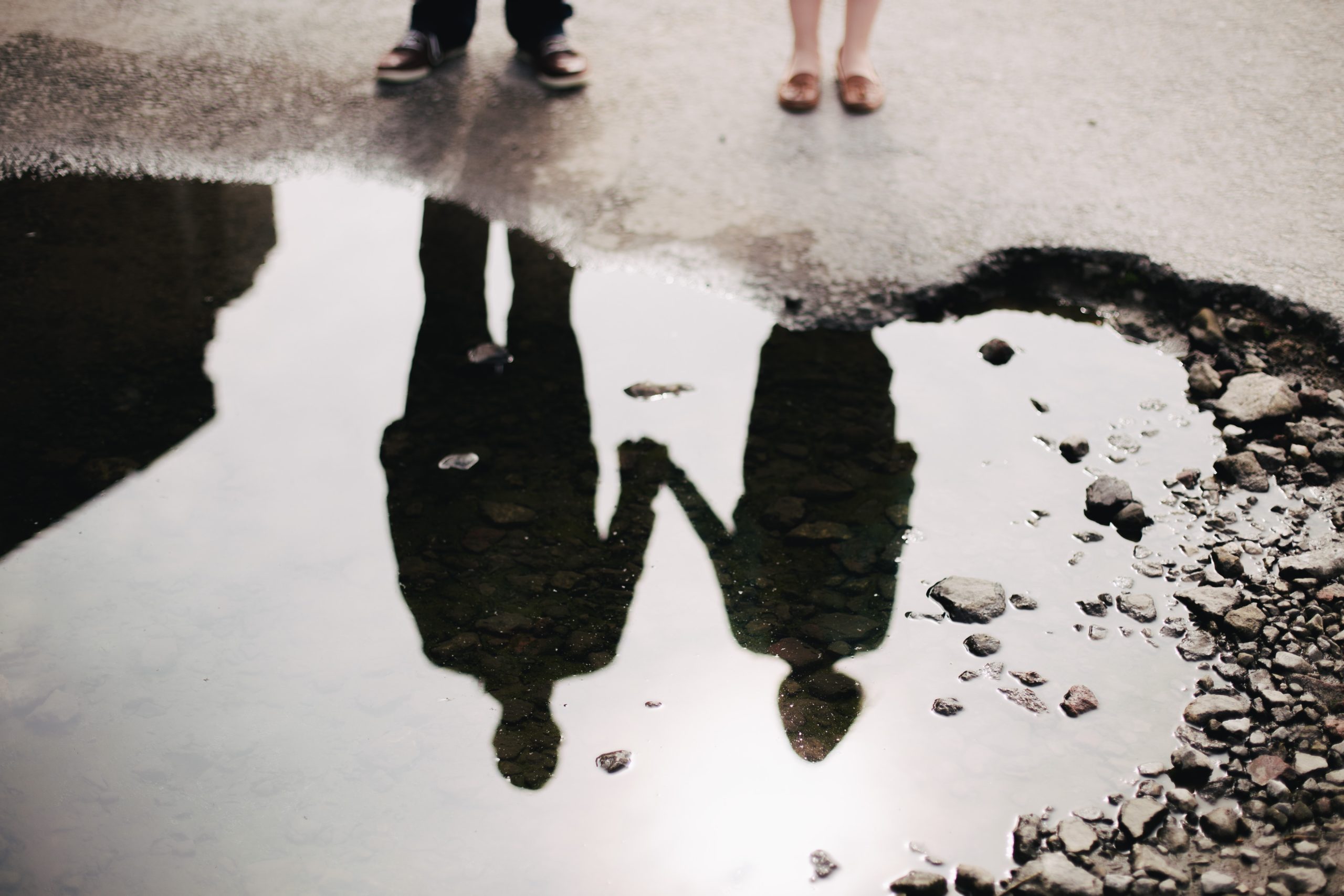 A couple standing next to each other, holding hands, reflected in a puddle.