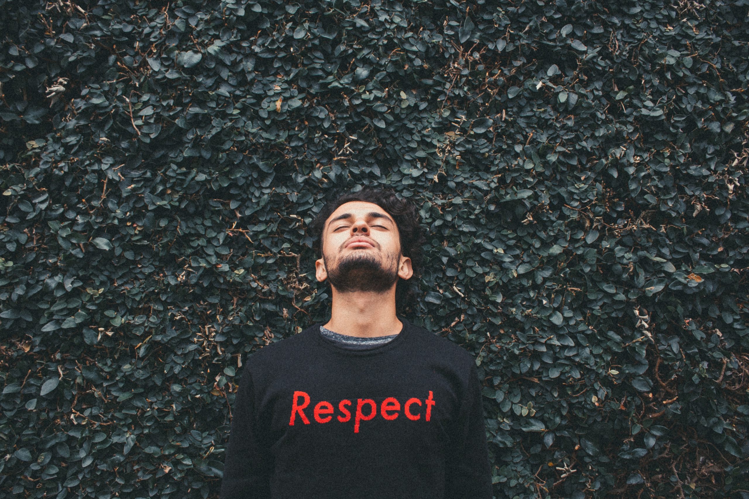A man standing in front of a hedge, looking up, wearing a black sweater with the word 'respect' on it.