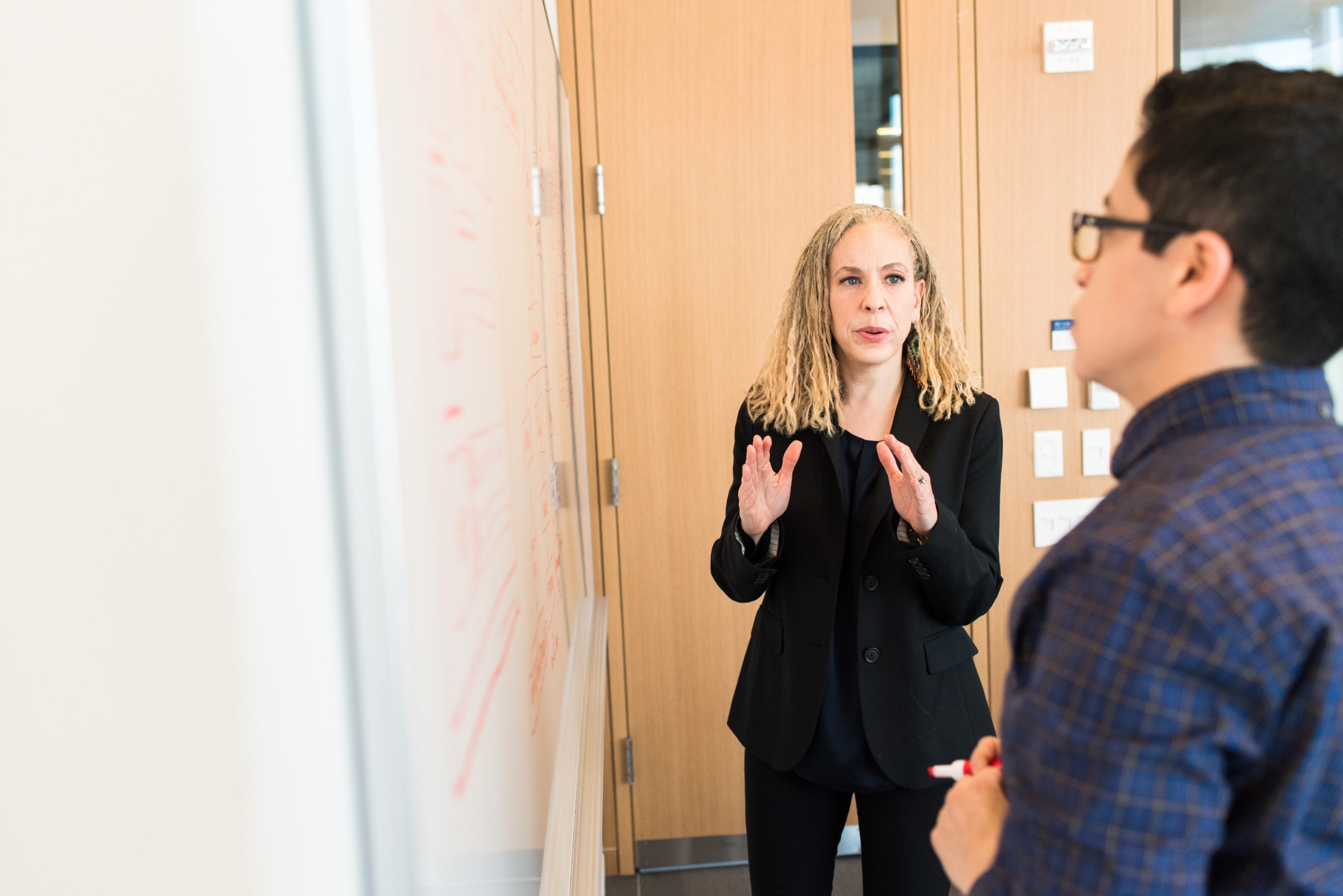 A woman speaking to a man, both standing in front of a white board.