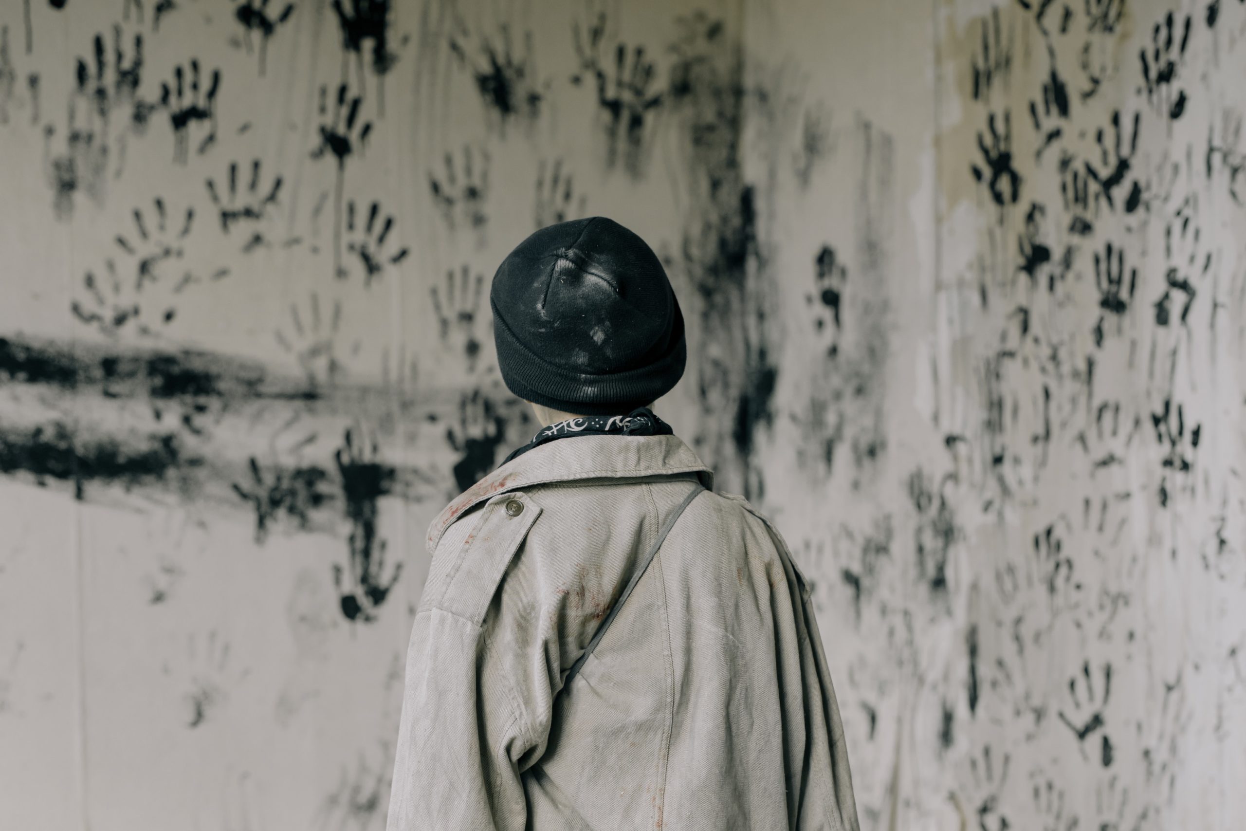 Someone in a beige jacket and black beanie facing a white wall that's covered in black handprints.