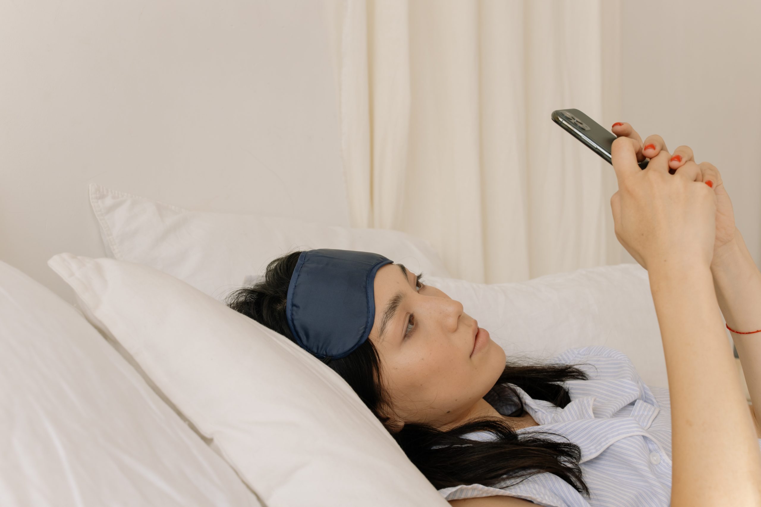 A woman laying in bed, typing on her phone, an eyemask pushed up onto her forehead.