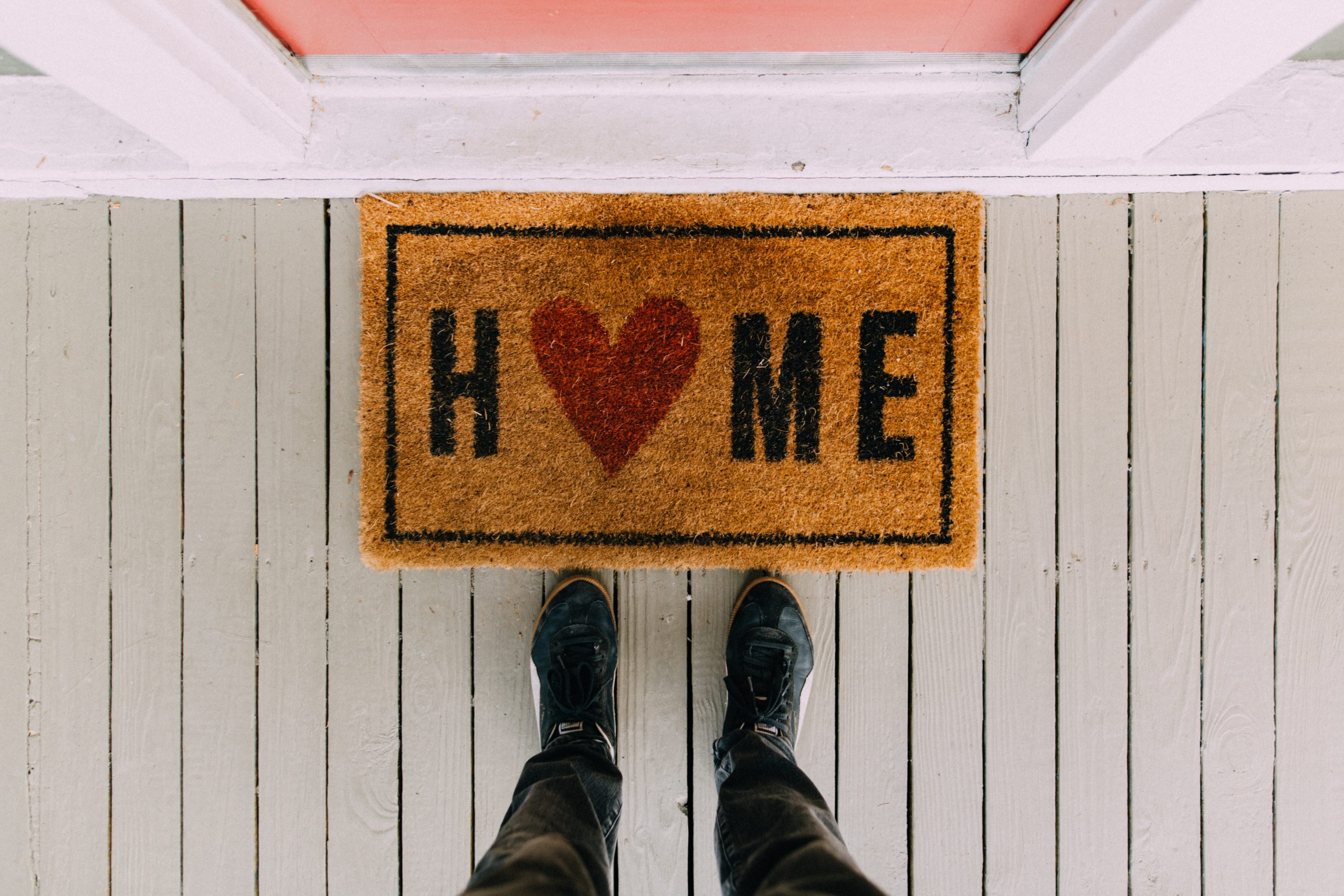 Someone standing in front of a doormat that says 'HOME' in black letters, save for the O, which is a red heart.