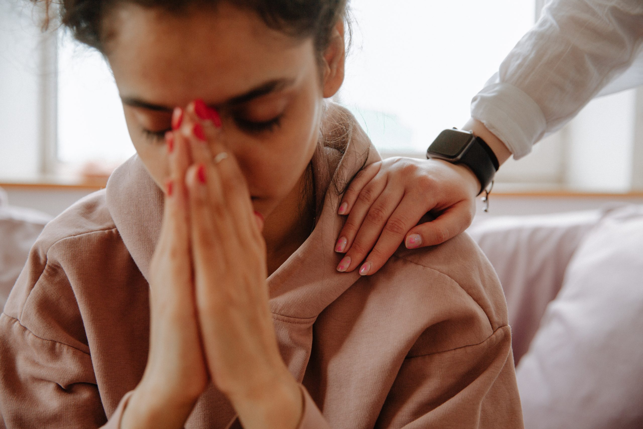 A woman with her hands together, pressed to her face, her friend's hand on her shoulder in comfort.