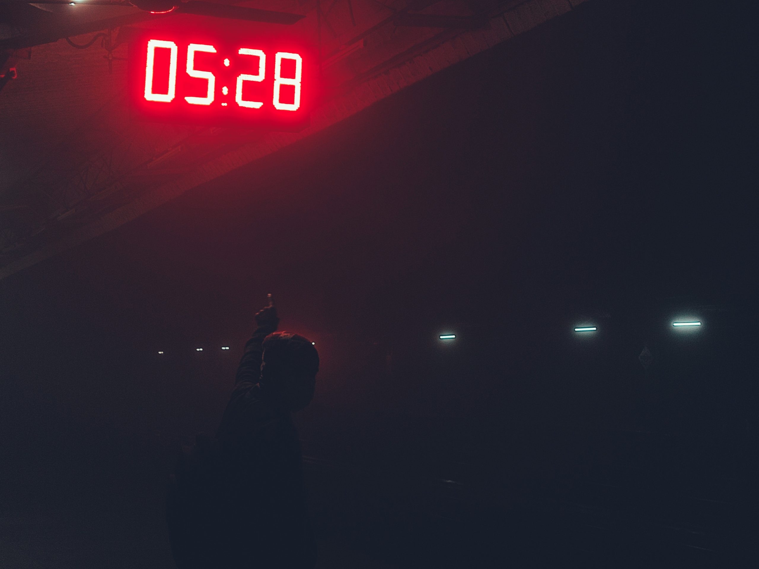 A red digital clock in the fog displaying 5:28, someone barely lit below pointing up to it.