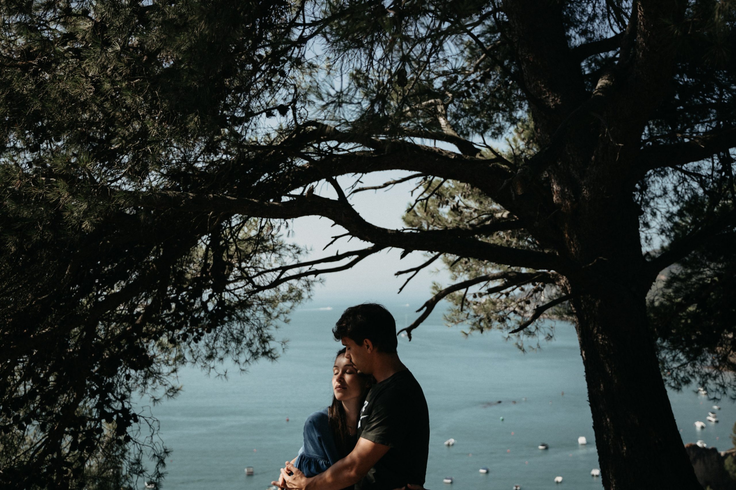 A couple embraces under a tree, in front of a lake.