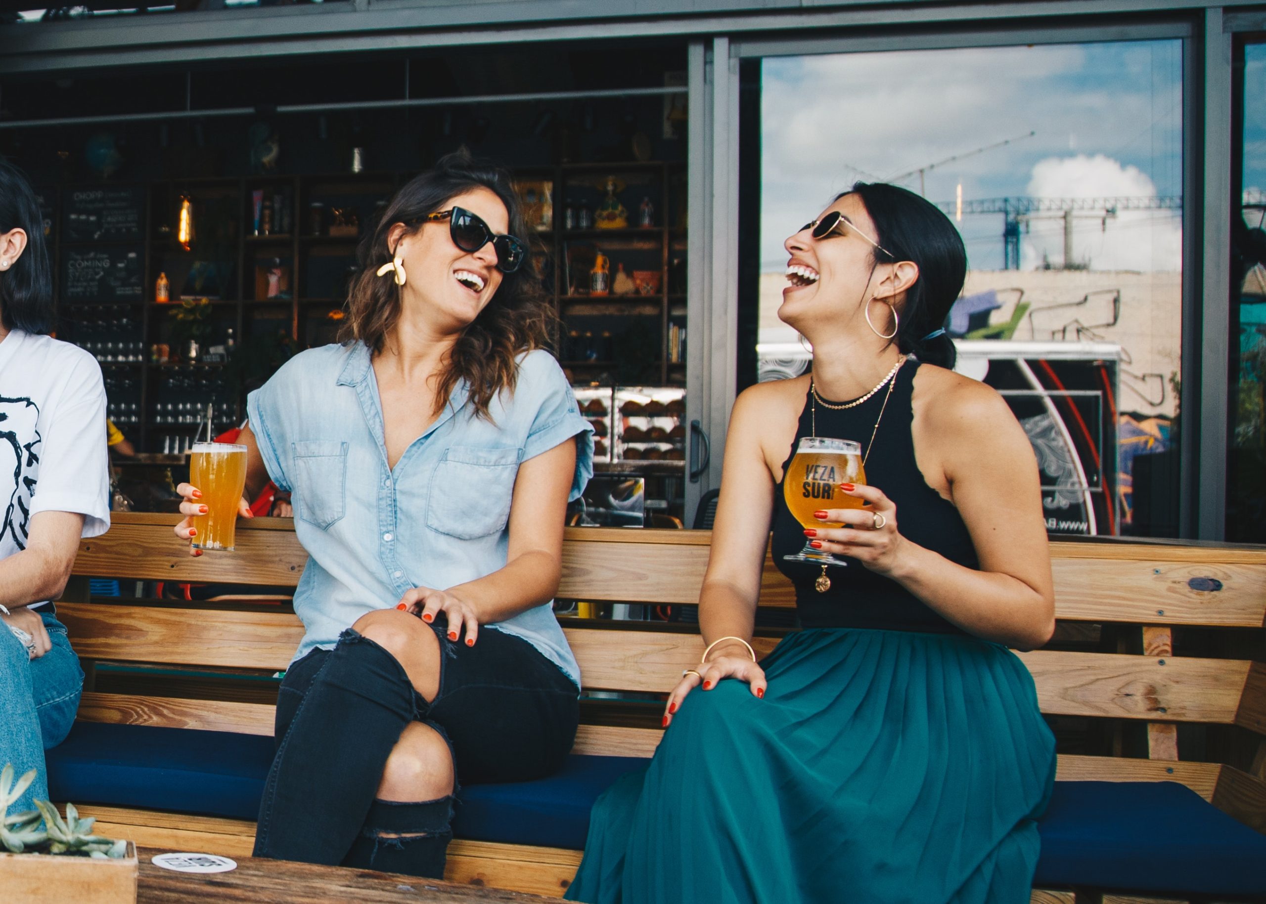 Two women sitting on a bench, one holding a drink, both laughing.