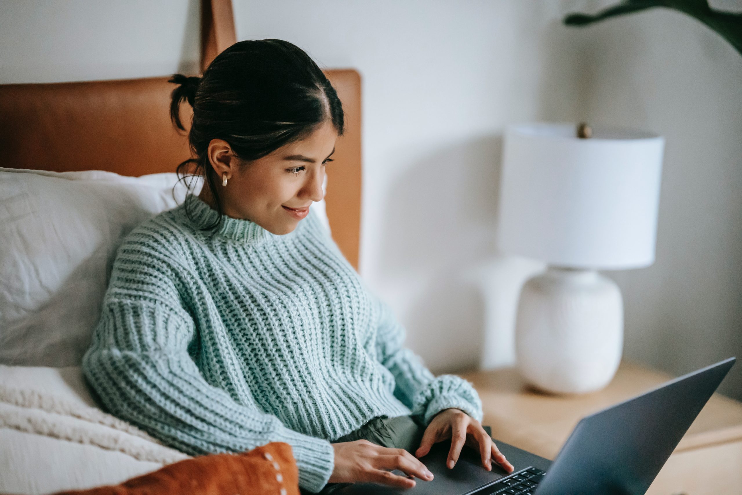 A woman sitting on her bed, typing on her laptop as she smiles.