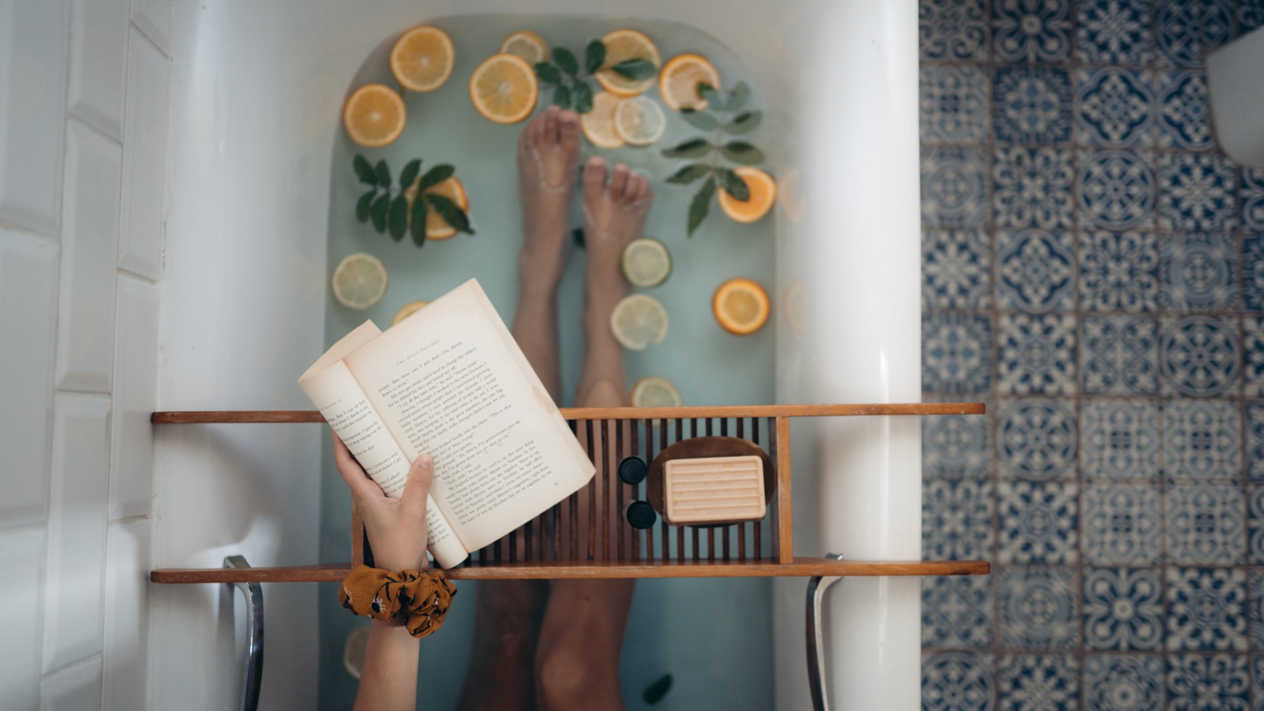 An arial shot of a woman in a bath, just her legs and arm visible. The water has lemon slices in it and she's reading a book on a wooden stand.