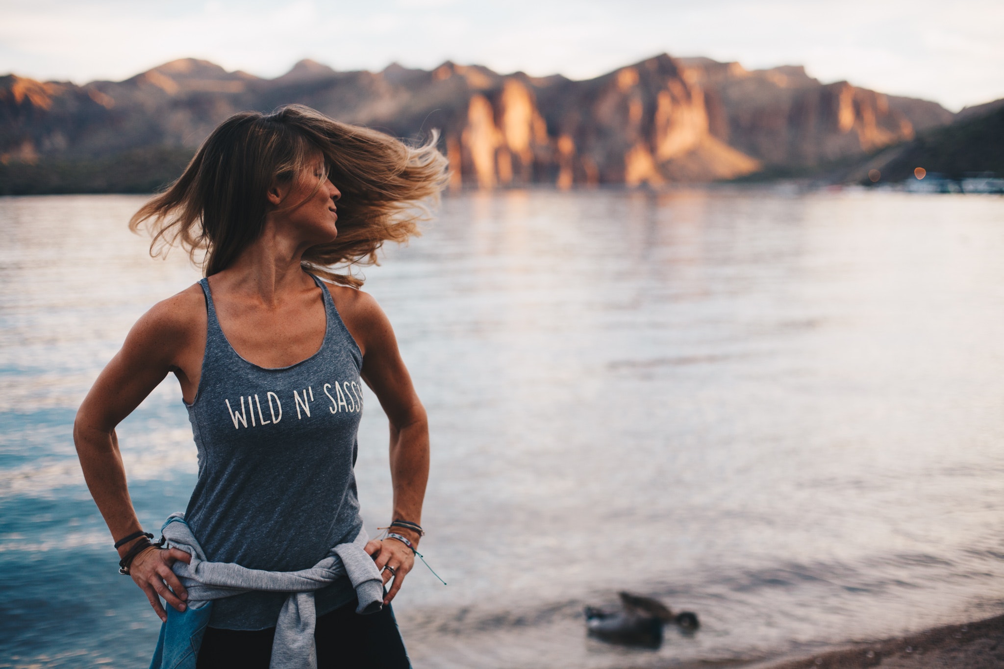 A woman standing in front of a small lake with her hands on her hips, shaking her hair around.