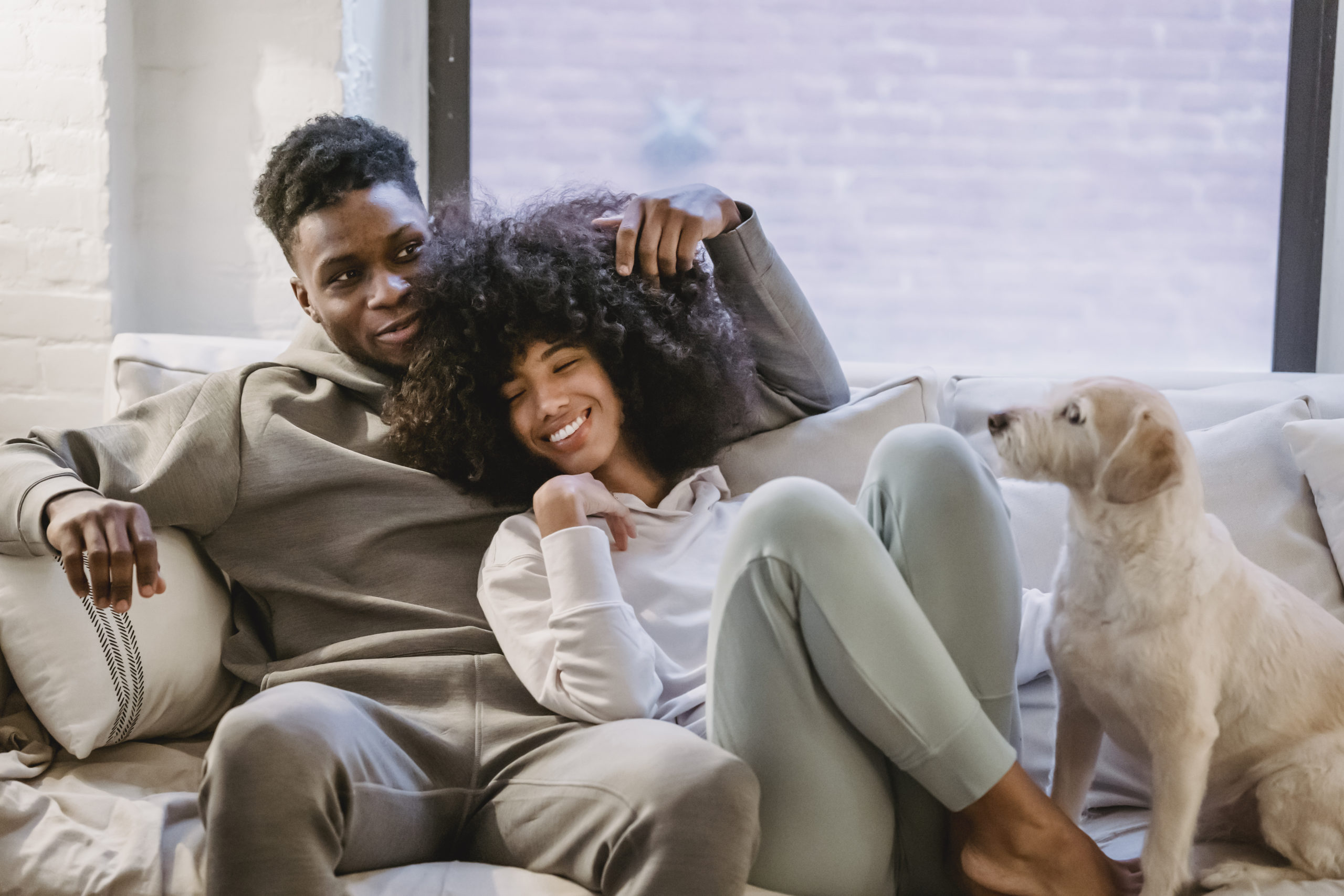 A couple sitting on a couch, the woman leaning into the man as she smiles, their dog next to them.