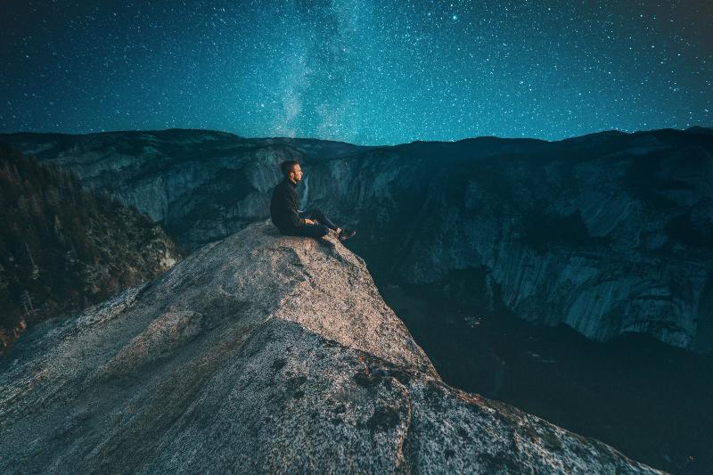man sits on cliff by starry blue sky