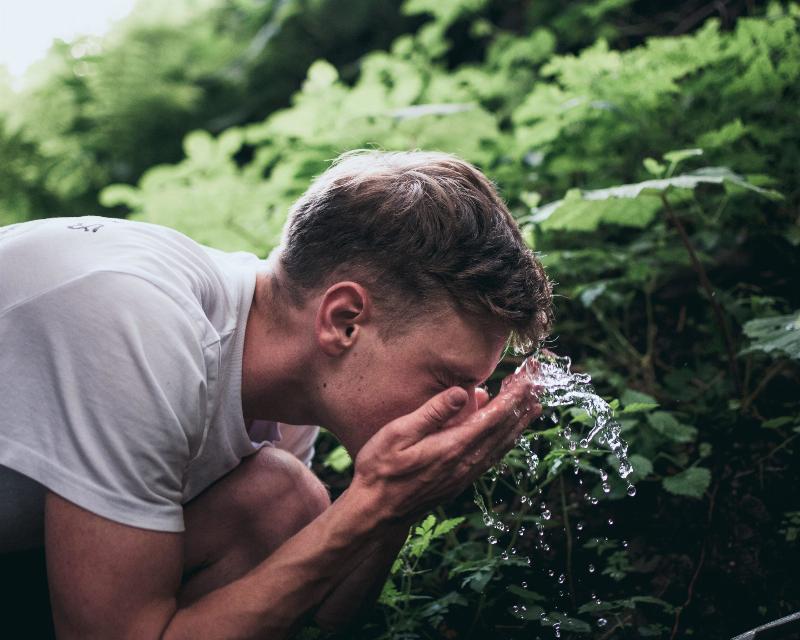 man splashes water on his face outdoors by plants