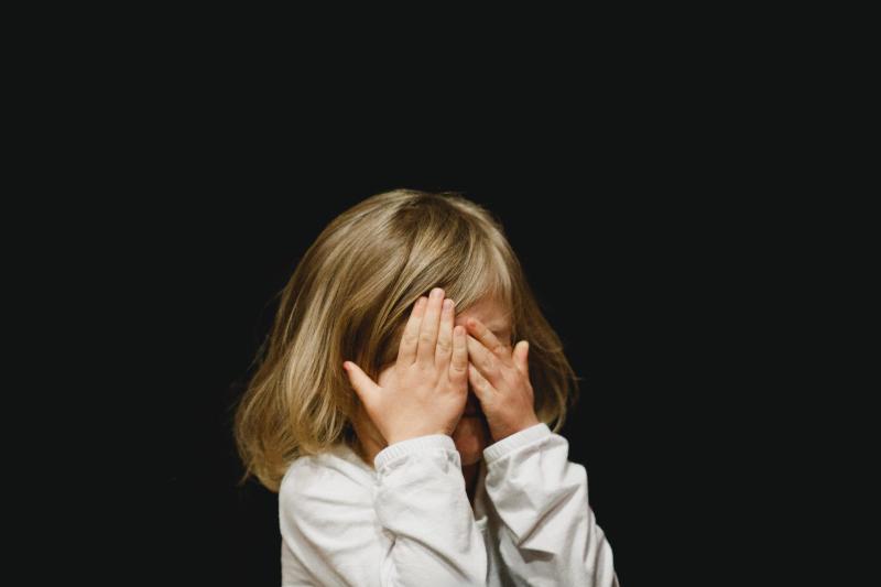 little girl hides her face with her hands on black background