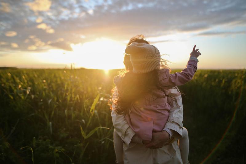 mom holds her daughter up in a field by the sun