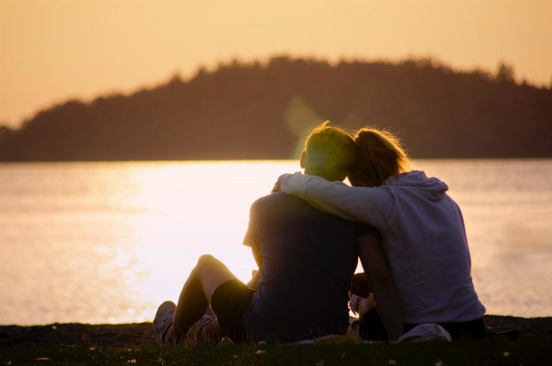 woman wraps her arm around man as they sit by the water in the sun reflection