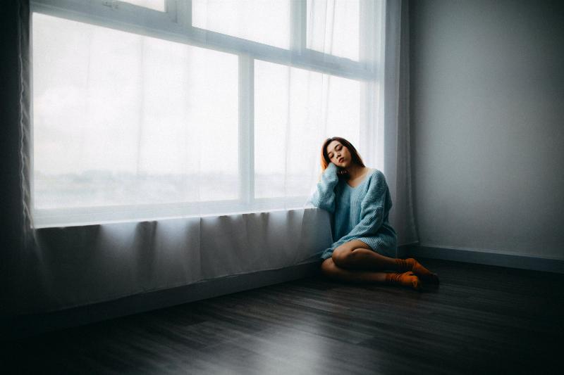 woman sits on the floor leaning by the window looking sad
