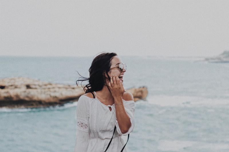 woman moves her hair away from face while smiling standing by the ocean