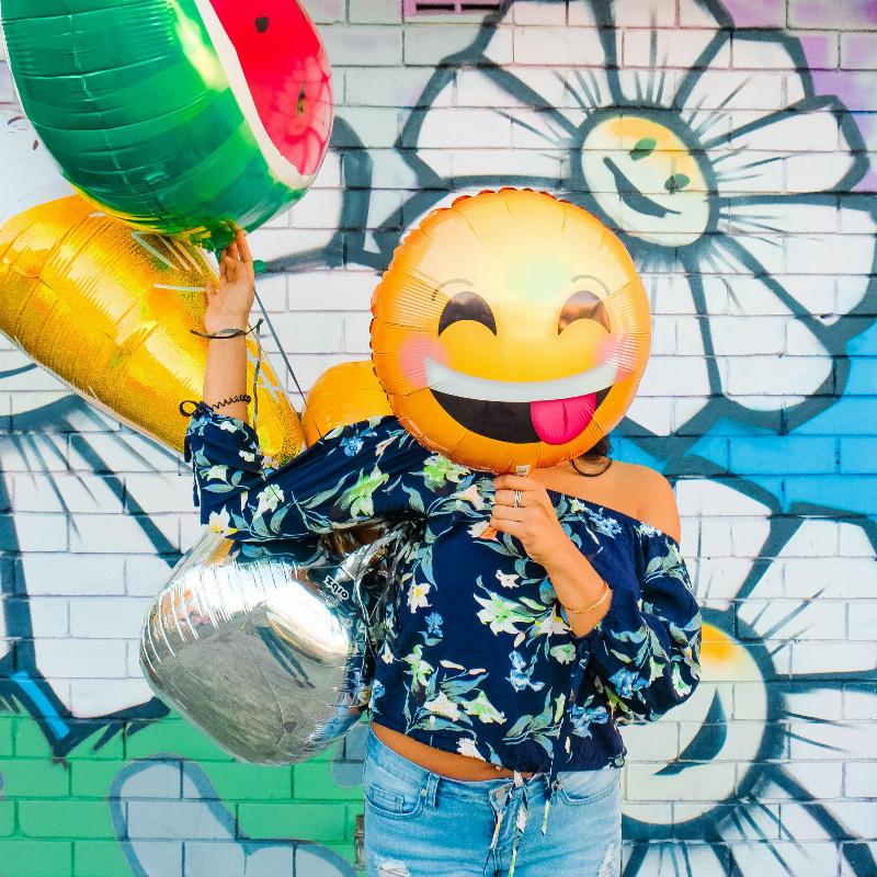 erson hides their face with smiley face balloon by colourful wall