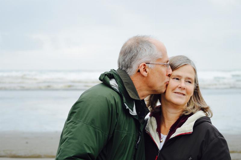 man kisses older woman on the cheek as they stand on the beach