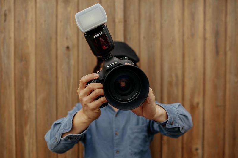 man holding up camera lens in button shirt by wooden wall