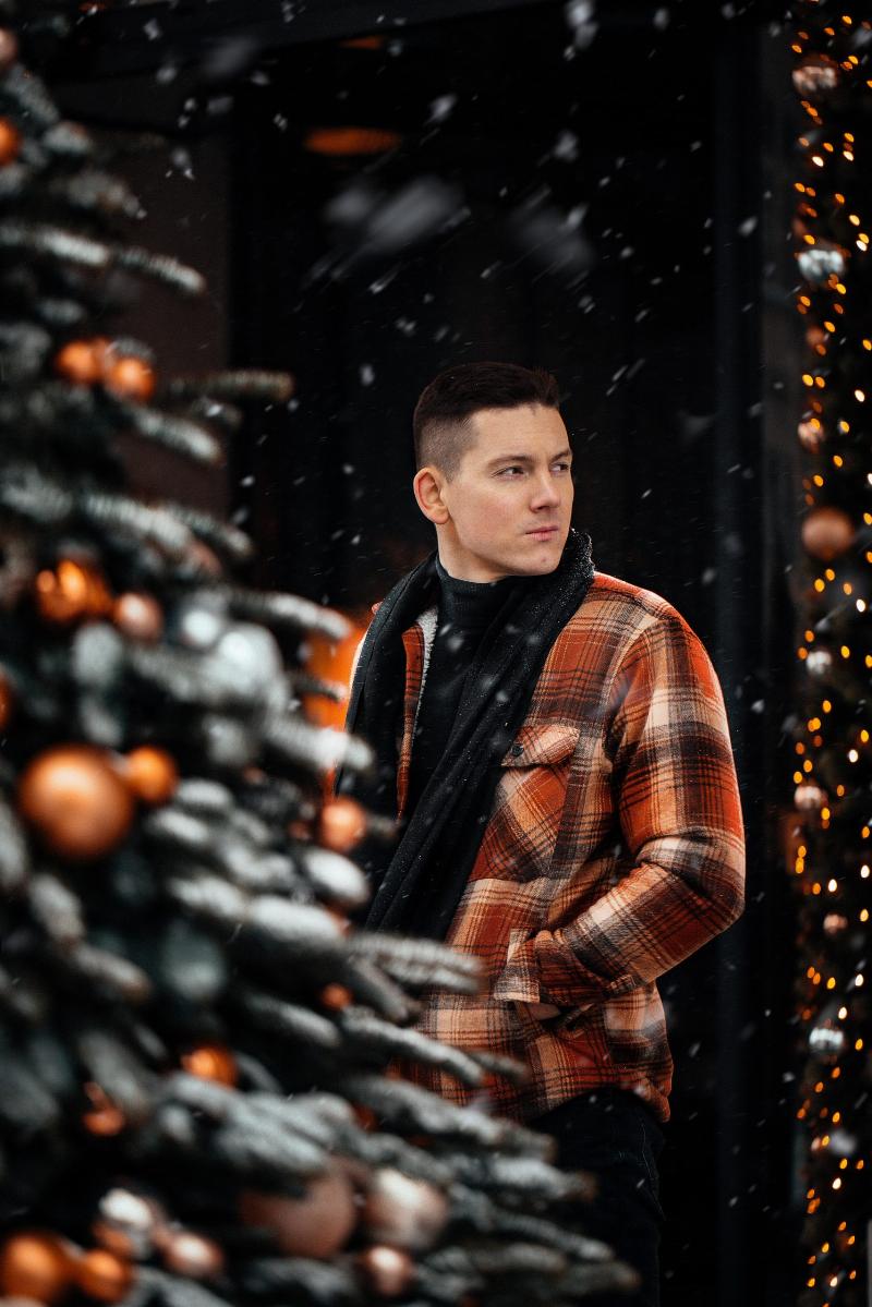 man stands behind christmas tree, look off to the side with his hand in his coat pocket