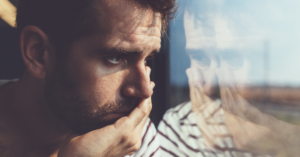 man looks out the window at his reflection looking sad with his hand on his face
