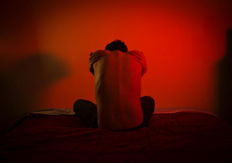 man hugs himself on bed in red lighting