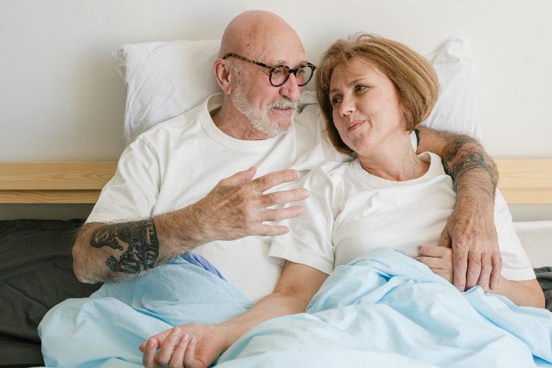 older man and woman sit in bed talking while wearing white tshirts