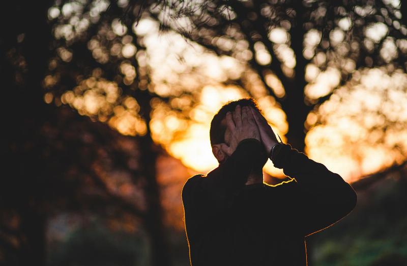 man covers his face with his hands while standing in sunset in stress