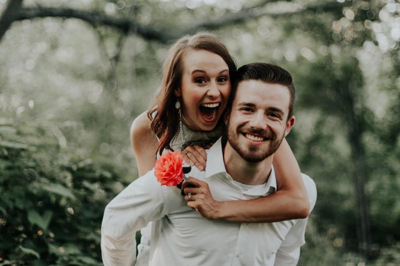 woman piggy backs on man's back while smiling and holding flower