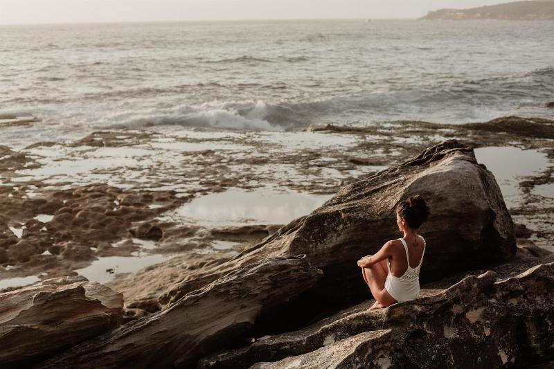woman sits on a rock facing teh cean
