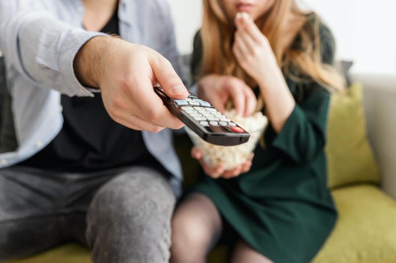 man puts remote up to tv with woman eating popcorn beside him