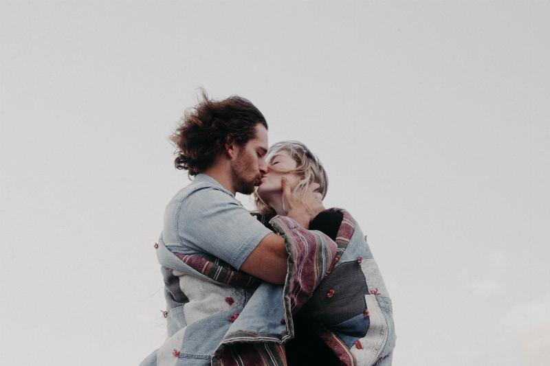 man holds woman's face as they kiss while wrapped in a blanket under a grey sky