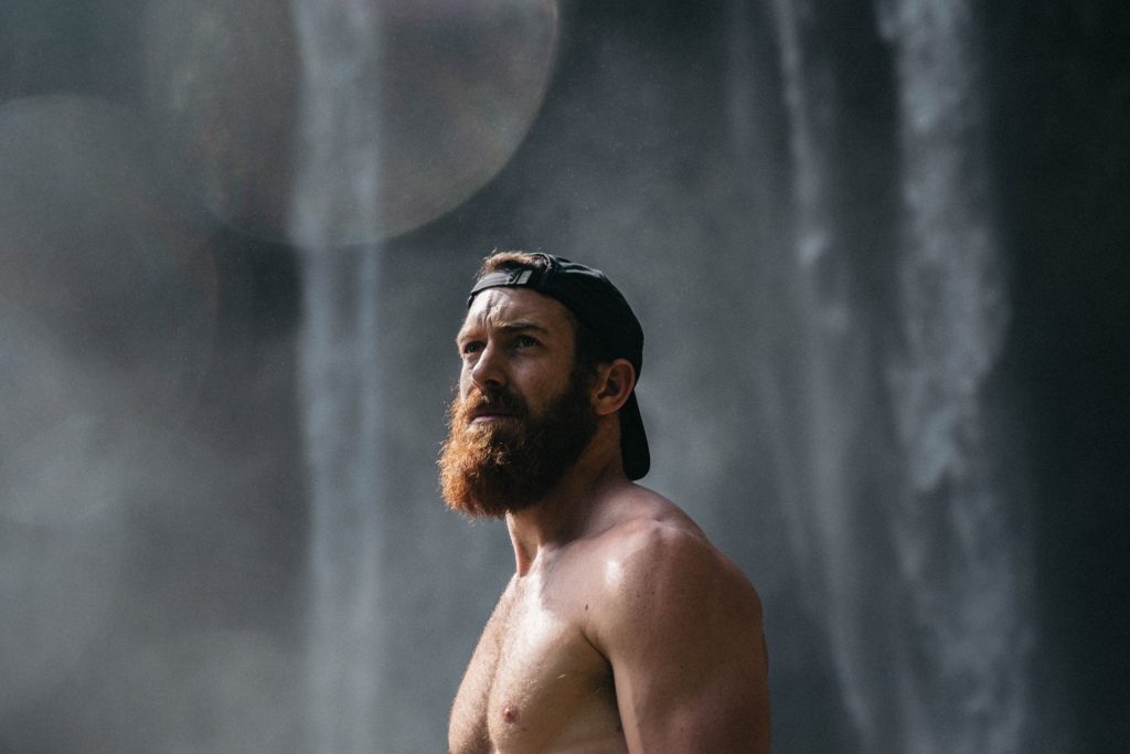 shirtless muscular man with hat stares into the distance by waterfall